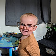 Albert is registered to the contest to win money with this photo: toddler, child, glasses, smile, indoor, table, toy, carpet, clothing, person, face, home, radiator, window_curtain, furniture, floor, playful, cute, young_child, portrait