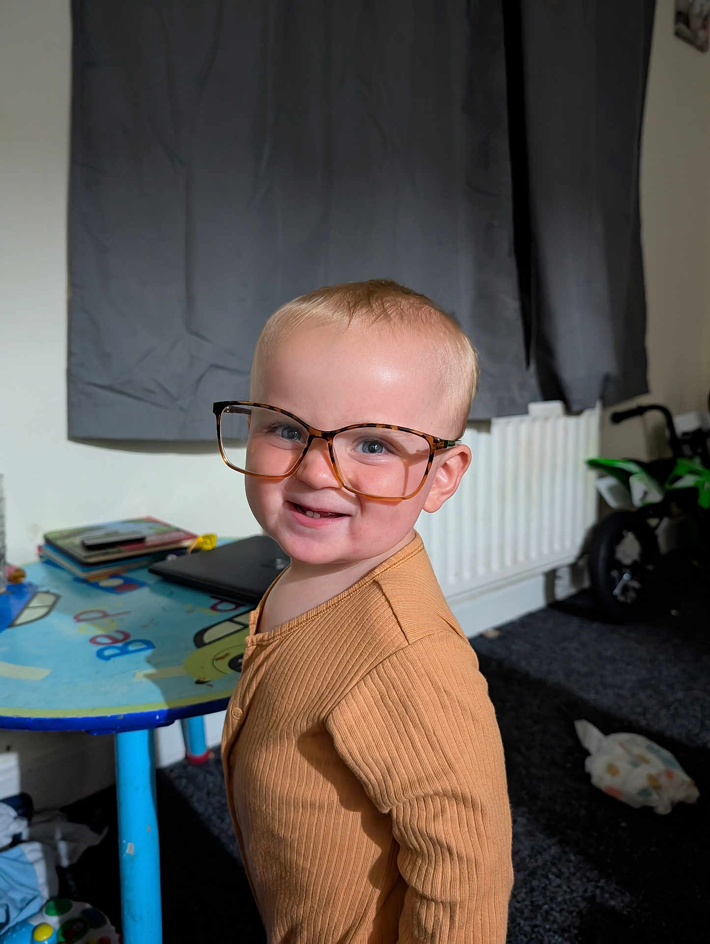 Albert is registered to the contest to win money with this photo: toddler, child, glasses, smile, indoor, table, toy, carpet, clothing, person, face, home, radiator, window_curtain, furniture, floor, playful, cute, young_child, portrait
