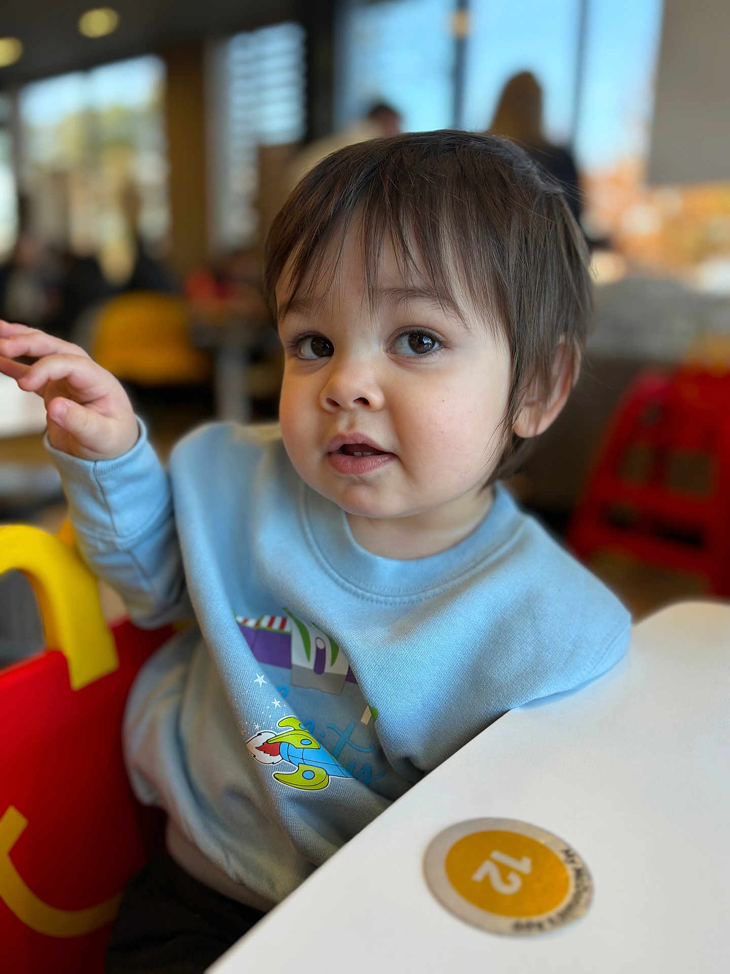 Fredrick joined the competition — help win amazing prizes! toddler, child, indoor, sitting, blue_sweatshirt, rocket_design, curious_expression, face, hair, table, chair, colorful, person, young_child, portrait, casual_clothing, daylight, blurry_background, cute, small_hand