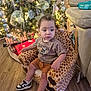 Aaron is registered to the contest to win money with this photo: toddler, child, plush_chair, giraffe_pattern, christmas_tree, ornaments, lights, presents, wood_floor, sneakers, shorts, tshirt, indoor, holiday, decorations, curious, seated, home, festive, cozy