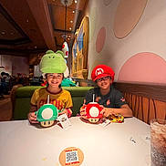 Kiara is registered to the contest to win money with this photo: child, hat, glasses, table, drink, restaurant, mushroom_cup, nintendo_world, smile, indoor, person, wall_decor, wood, lighting, fun, casual, food_and_drink, seat, tabletop, friends