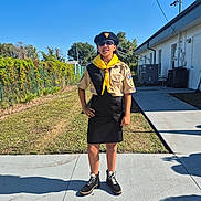 Kiara is registered to the contest to win money with this photo: child, scout_uniform, yellow_neckerchief, beret, sunglasses, outdoor, sidewalk, grass, building, shadow, tree, fence, person, boots, sunny, blue_sky, standing, hand_on_hip, concrete, patch_of_dirt