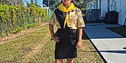 Kiara is registered to the contest to win money with this photo: child, scout_uniform, yellow_neckerchief, beret, sunglasses, outdoor, sidewalk, grass, building, shadow, tree, fence, person, boots, sunny, blue_sky, standing, hand_on_hip, concrete, patch_of_dirt