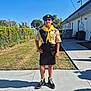 child, scout_uniform, yellow_neckerchief, beret, sunglasses, outdoor, sidewalk, grass, building, shadow, tree, fence, person, boots, sunny, blue_sky, standing, hand_on_hip, concrete, patch_of_dirt