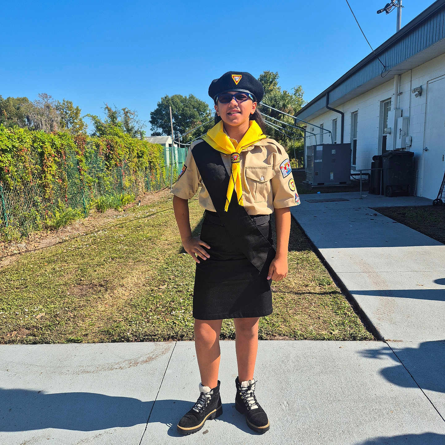 Kiara is registered to the contest to win money with this photo: beret, blue_sky, boots, building, child, concrete, fence, grass, hand_on_hip, outdoor, patch_of_dirt, person, scout_uniform, shadow, sidewalk, standing, sunglasses, sunny, tree, yellow_neckerchief