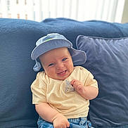 Cooper is registered to the contest to win money with this photo: baby, smiling, hat, blue_hat, cream_shirt, denim_shorts, couch, pillow, indoor, happy, child, infant, seated, cute, portrait, soft_light, cozy, casual_clothing, person, relaxed