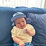baby, smiling, hat, blue_hat, cream_shirt, denim_shorts, couch, pillow, indoor, happy, child, infant, seated, cute, portrait, soft_light, cozy, casual_clothing, person, relaxed