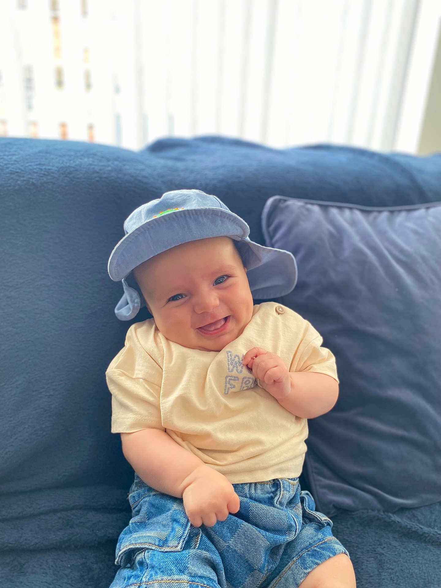 Cooper is registered to the contest to win money with this photo: baby, smiling, hat, blue_hat, cream_shirt, denim_shorts, couch, pillow, indoor, happy, child, infant, seated, cute, portrait, soft_light, cozy, casual_clothing, person, relaxed