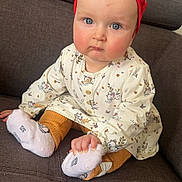 élia a rejoint le concours — aidez-le/la à gagner de superbes lots ! baby, child, infant, sitting, couch, red_headband, blue_eyes, white_dress, patterned_clothing, orange_pants, white_socks, hand, face, cute, indoors, furniture, portrait, expression, small_child, young