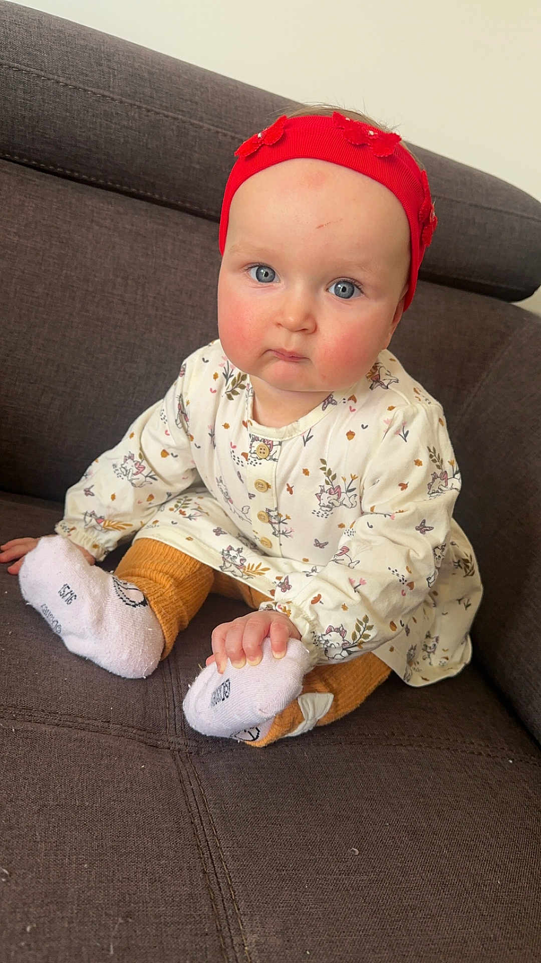 élia a rejoint le concours — aidez-le/la à gagner de superbes lots ! baby, child, infant, sitting, couch, red_headband, blue_eyes, white_dress, patterned_clothing, orange_pants, white_socks, hand, face, cute, indoors, furniture, portrait, expression, small_child, young