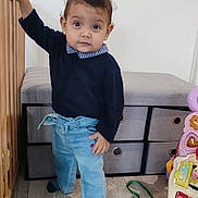 Maria a rejoint le concours — aidez-le/la à gagner de superbes lots ! toddler, child, toy, wooden_crib, storage_bins, floor, shirt, pants, sock, indoor, playroom, curly_hair, earrings, striped_socks, blue_pants, black_sweater, wood_floor, colorful_toys, standing, curious