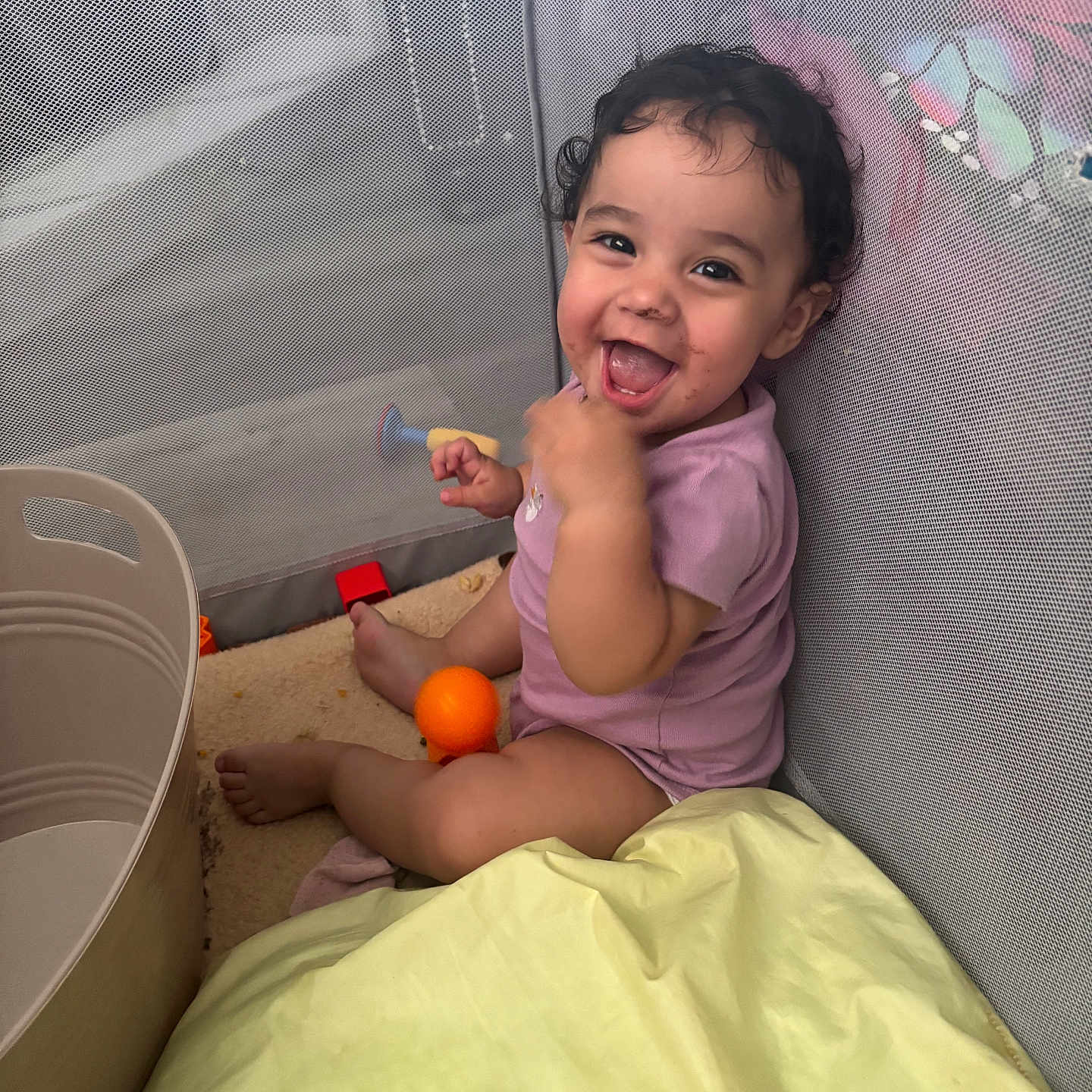 Pet is registered to the contest to win money with this photo: baby, child, childhood, curly_hair, cushion, cute, face, fun, happy, indoor, infant, orange_ball, person, play, playpen, purple_onesie, sitting, smiling, toy, young