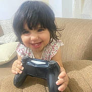 Mariam joined the competition — help win amazing prizes! toddler, child, smile, gaming_controller, couch, sofa, indoor, person, young_child, happy, hand, hair, clothing, floral_pattern, furniture, living_room, pillow, cute, playful, relaxing