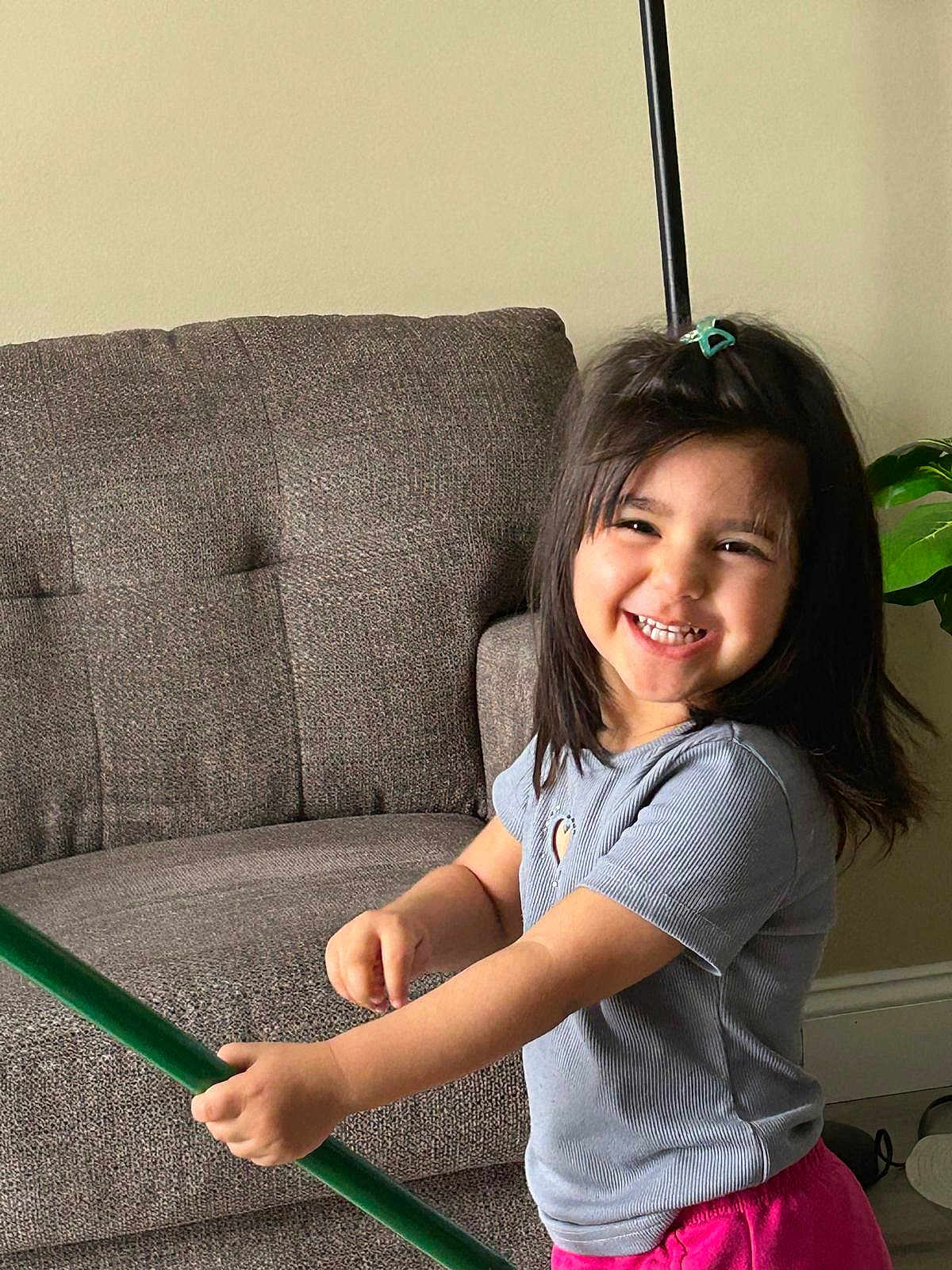 Mariam is registered to the contest to win money with this photo: child, girl, smiling, happy, indoor, couch, furniture, plant, green_stick, hair_clip, short_sleeve_shirt, pink_pants, living_room, playful, cute, portrait, person, home, fun, young
