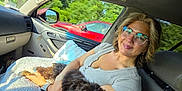 Smudge joined the competition — help win amazing prizes! woman, dog, glasses, car_interior, seat, window, pillow, sunlight, smile, cuddling, pet, travel, nature, greenery, red_car, casual_clothing, human, comfort, happy, relaxation
