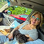 woman, dog, glasses, car_interior, seat, window, pillow, sunlight, smile, cuddling, pet, travel, nature, greenery, red_car, casual_clothing, human, comfort, happy, relaxation