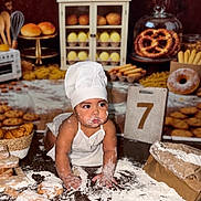 Gabriel is registered to the contest to win money with this photo: baby, child, infant, chef_hat, bakery, baking, bread, pastry, pretzel, cookies, flour, messy, kitchen, food, sitting, portrait, costume, white_clothing, rolling_pin, birthday_photo