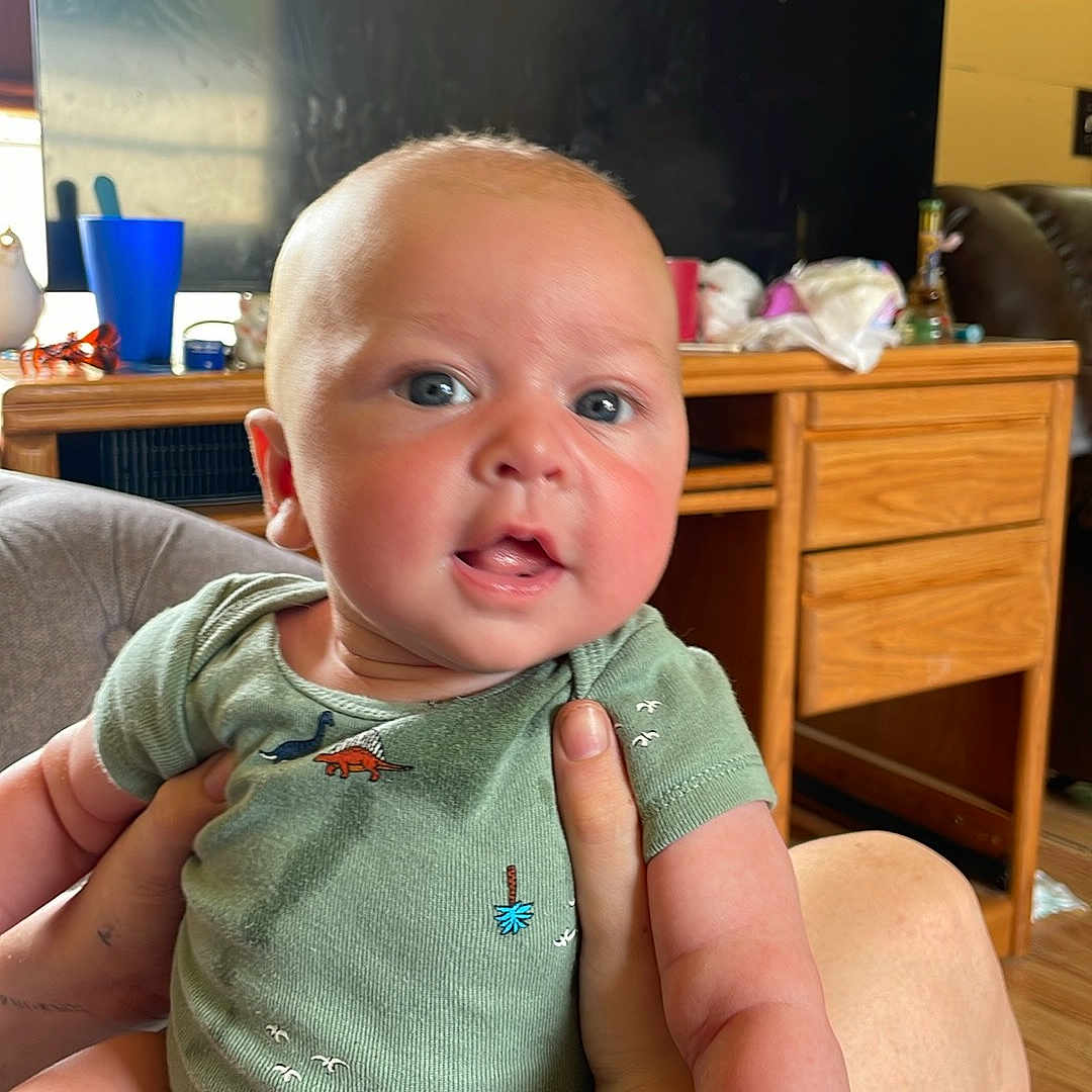 Grayson is registered to the contest to win money with this photo: baby, blue_eyes, bright, candid, child, cute, desk, dinosaur_print, expression, face, furniture, hand, holding, home, indoor, infant, onesie, person, skin, smiling