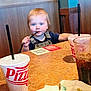 Axel is registered to the contest to win money with this photo: toddler, child, blue_eyes, blonde_hair, straw, table, restaurant, pizza, cup, drink, napkin, wooden_bench, casual, indoor, person, smiling, playful, clothing, overalls, dining