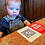 toddler, child, blue_eyes, overalls, striped_clothing, high_chair, restaurant, table, placard, qr_code, pizza_ranch, face, curious, messy, food, indoor, wood_paneling, person, clothing, seat