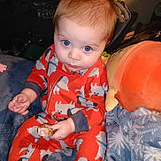 Axel is registered to the contest to win money with this photo: baby, child, onesie, orange_clothing, dog_prints, blue_eyes, plush_toy, blanket, food, sitting, indoor, cute, curious, infant, portrait, hand, furniture, soft_toy, cozy, expression