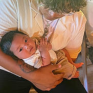 Keano John is registered to the contest to win money with this photo: baby, adult, holding, arms, indoor, clothing, curly_hair, glasses, necklaces, tattoo, skin, person, seated, shirt, pants, foot, hand, looking, gentle, warm_light