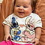 baby, child, smiling, happy, tshirt, pink_shorts, cushion, brown_background, cute, infant, portrait, indoors, chubby_cheeks, sitting, clothing, person, cute_expression, shorts, soft_texture, joy