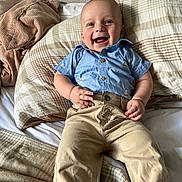 Vinny is registered to the contest to win money with this photo: baby, child, smiling, blue_shirt, beige_pants, bed, blanket, pillow, happy, infant, person, portrait, indoors, clothing, cute, sitting, human, face, skin, young