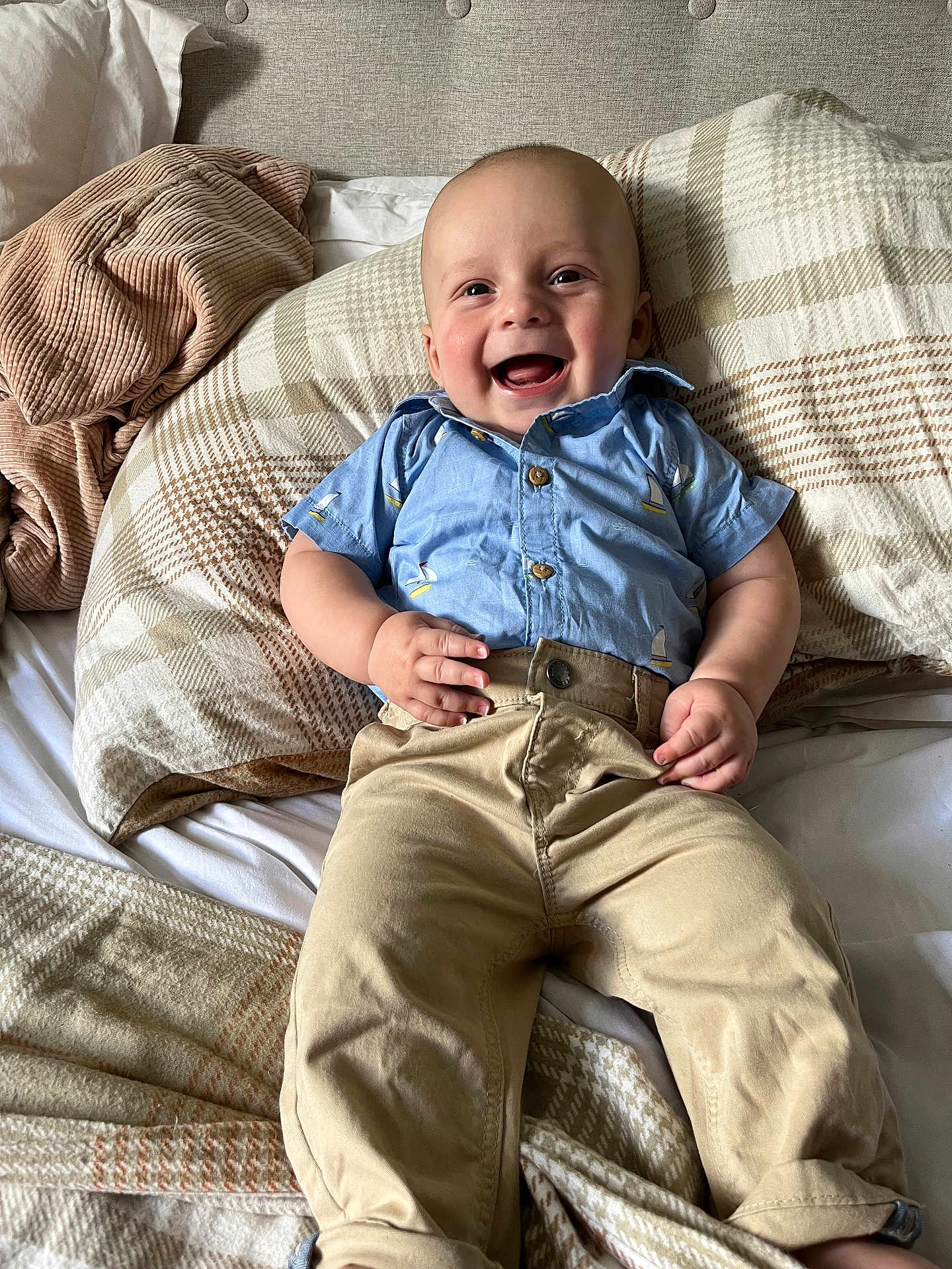 Vinny is registered to the contest to win money with this photo: baby, child, smiling, blue_shirt, beige_pants, bed, blanket, pillow, happy, infant, person, portrait, indoors, clothing, cute, sitting, human, face, skin, young