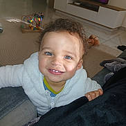 Rayan participe au concours pour gagner de l'argent avec cette photo : toddler, child, smiling, curly_hair, blue_eyes, white_jacket, indoors, couch, toy, bead_maze, television, carpet, floor, living_room, furniture, person, happy, portrait, playful, cute