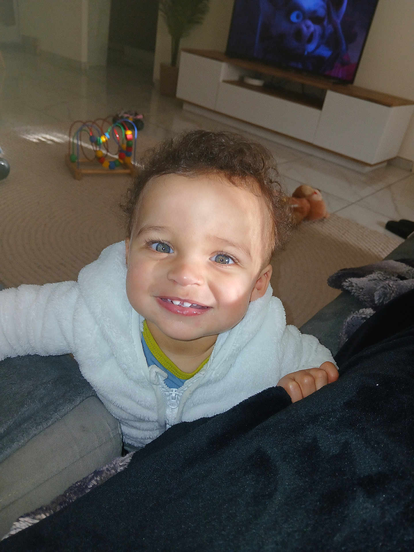 Rayan participe au concours pour gagner de l'argent avec cette photo : toddler, child, smiling, curly_hair, blue_eyes, white_jacket, indoors, couch, toy, bead_maze, television, carpet, floor, living_room, furniture, person, happy, portrait, playful, cute