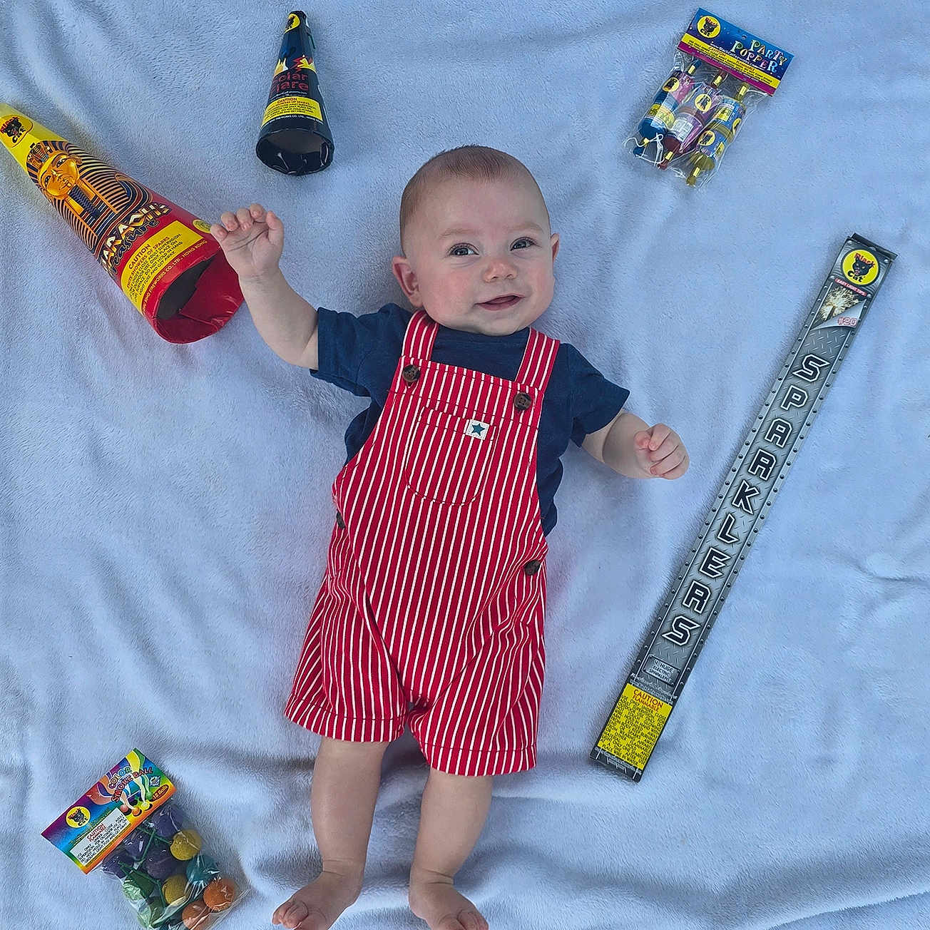 Revan is registered to the contest to win money with this photo: baby, blanket, blue_shirt, celebration, child, colorful, cute, footwearless, happy, indoors, infant, lying_down, party, party_poppers, playful, red_striped_overalls, small_hands, smile, soft_texture, sparklers