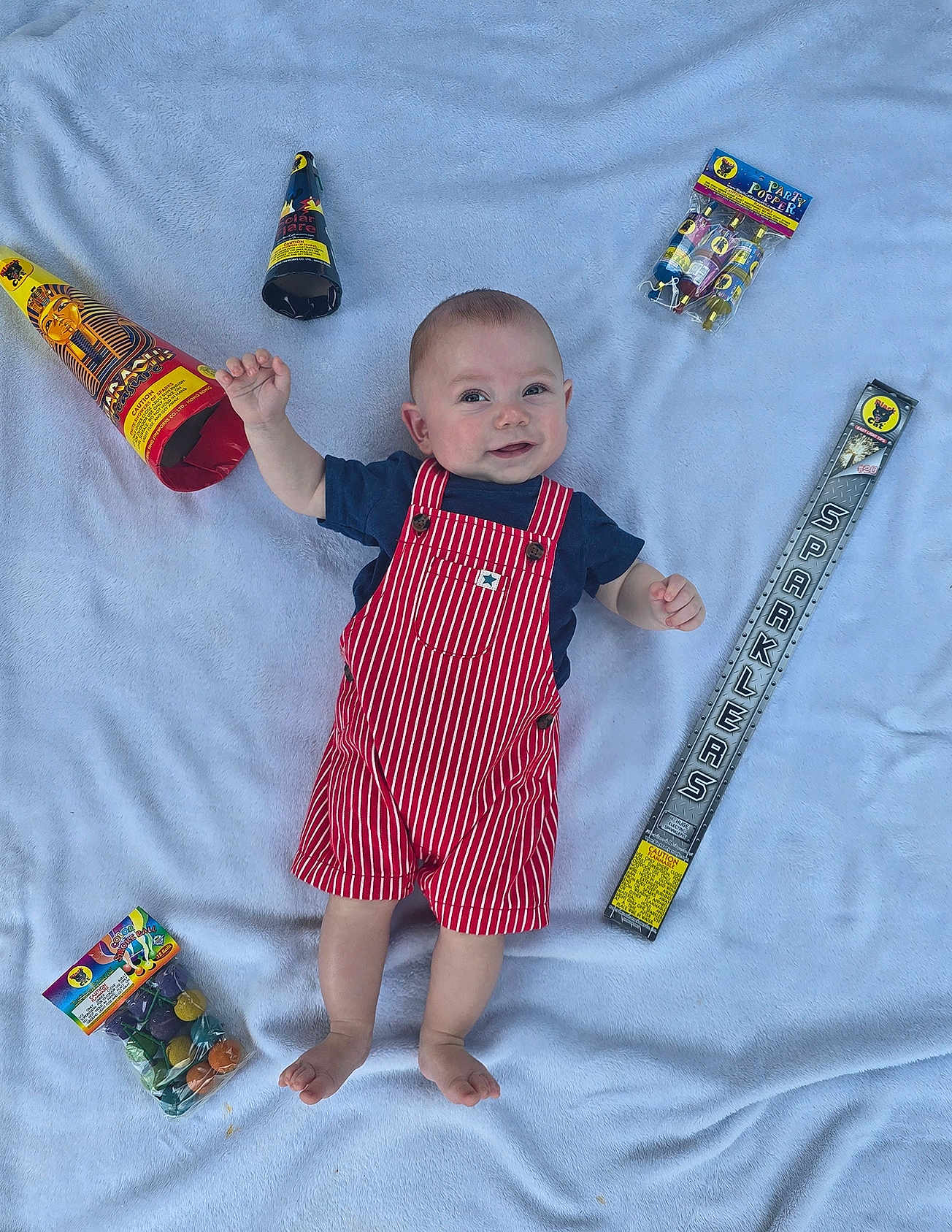 Revan is registered to the contest to win money with this photo: baby, infant, child, red_striped_overalls, blue_shirt, party_poppers, sparklers, blanket, smile, happy, celebration, colorful, footwearless, lying_down, cute, party, small_hands, soft_texture, indoors, playful