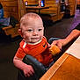 Revan is registered to the contest to win money with this photo: baby, child, smiling, orange_shirt, high_chair, bread_roll, restaurant, wooden_booth, table, hand, adult, indoor, cute, face, person, casual_clothing, dining, food, seat, happy
