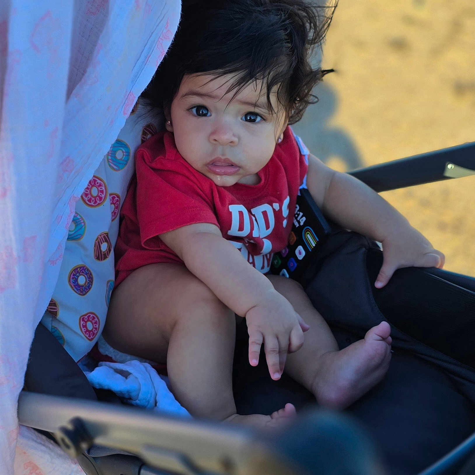 Juliett joined the competition — help win amazing prizes! baby, child, stroller, red_shirt, donut_pattern, blanket, curly_hair, outdoor, sunlight, infant, face, portrait, cute, seated, foot, hand, clothing, expression, baby_gear, soft_light