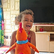 Sarah participe au concours pour gagner de l'argent avec cette photo : bird, cabinet, child, colorful, curly_hair, curtains, furniture, happy, indoor, laughing, living_room, orange, person, playful, plush_toy, smiling, stone_wall, table, television, young