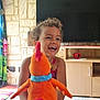 bird, cabinet, child, colorful, curly_hair, curtains, furniture, happy, indoor, laughing, living_room, orange, person, playful, plush_toy, smiling, stone_wall, table, television, young