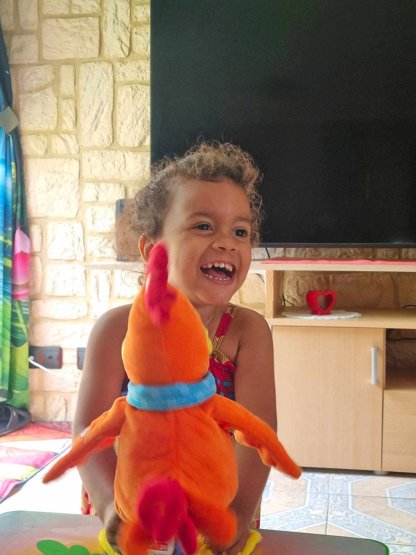 Sarah participe au concours pour gagner de l'argent avec cette photo : bird, cabinet, child, colorful, curly_hair, curtains, furniture, happy, indoor, laughing, living_room, orange, person, playful, plush_toy, smiling, stone_wall, table, television, young