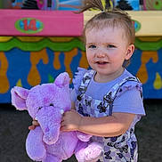 Rylee is registered to the contest to win money with this photo: toddler, child, plush_toy, purple_elephant, smiling, outdoor, carnival, colorful_background, short_hair, pony_tail, overalls, butterfly_pattern, holding_toy, daylight, cute, happy, person, face, playful, fun