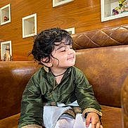 Garvith joined the competition — help win amazing prizes! toddler, child, traditional_clothing, green_shirt, white_socks, brown_couch, curly_hair, smiling, indoor, wooden_wall, decorative_shelves, figurines, cute, sitting, portrait, young_child, happy, cozy, home_interior, playful