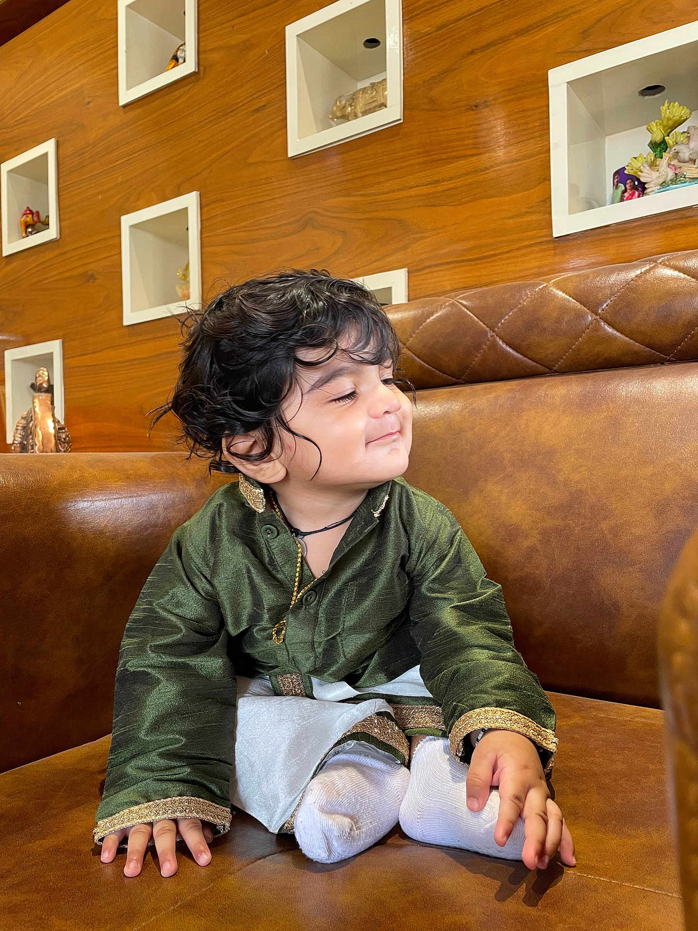 Garvith joined the competition — help win amazing prizes! toddler, child, traditional_clothing, green_shirt, white_socks, brown_couch, curly_hair, smiling, indoor, wooden_wall, decorative_shelves, figurines, cute, sitting, portrait, young_child, happy, cozy, home_interior, playful