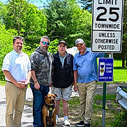 Sarge is registered to the contest to win money with this photo: man, dog, road_sign, outdoor, trees, sidewalk, grass, casual_clothing, smile, sunglasses, cap, pavement, group, leash, sunny, nature, greenery, street, walking_path, people_standing
