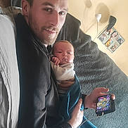 Noé a rejoint le concours — aidez-le/la à gagner de superbes lots ! man, father, baby, infant, couch, blue_blanket, phone, charger, wall_art, painting, lamp, pillow, beard, holding, indoor, portrait, smartphone_screen, relaxing, parenting, home