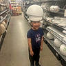 aisle, black_pants, blue_shoes, boxes, boy, casual_clothing, child, floor, fun, headwear, helmet, home_goods, indoor, person, retail, shelves, shopping, smiling, store, styrofoam