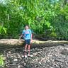 adventure, boy, casual_clothing, child, daylight, environment, forest, greenery, leaves, nature, outdoor, playful, rocks, shirt, shoes, shorts, standing, summer, sunlight, tree_trunk