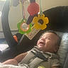 baby, baby_toy, blanket, car_seat, child, childcare, clothing, face, flower_toy, hanging_toys, happy, head, indoor, infant, portrait, seatbelt, smiling, toy, window_light, young_child