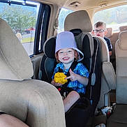Legend is registered to the contest to win money with this photo: child, toddler, car_seat, bucket_hat, toy, glasses, car_interior, seatbelt, smile, short_hair, window, daylight, vehicle, seat, blue_shirt, plaid, person, safety, happy, family