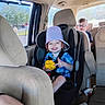 child, toddler, car_seat, bucket_hat, toy, glasses, car_interior, seatbelt, smile, short_hair, window, daylight, vehicle, seat, blue_shirt, plaid, person, safety, happy, family