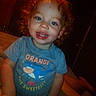 child, toddler, curly_hair, blue_eyes, smiling, shirt, tshirt, text_on_clothes, indoor, bed, wooden_door, door_handle, person, happy, cute, face, portrait, young_child, sitting, playful