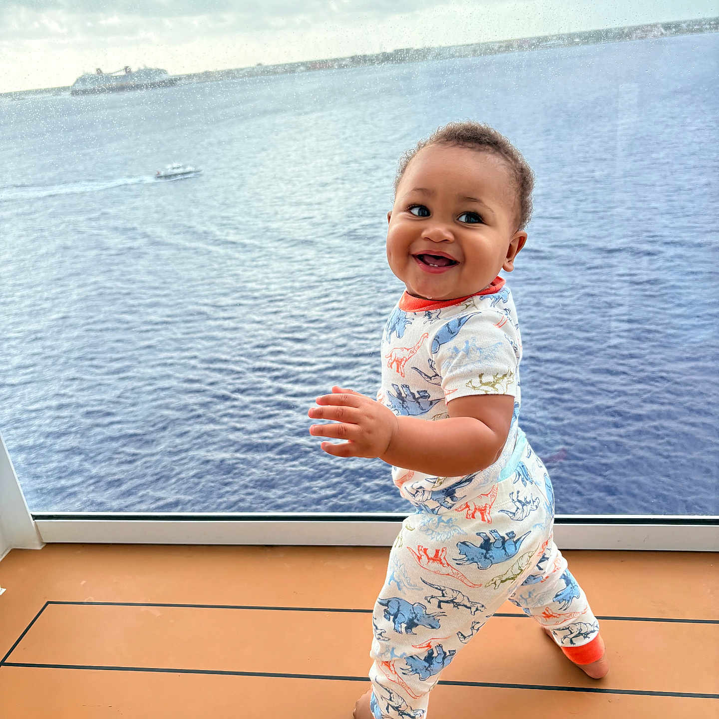 Laviarus is registered to the contest to win money with this photo: apparel, baby, child, cloudy, cute, daylight, deck, dinosaur_pajamas, footwear, happy, ocean, outdoor, person, playful, sky, smiling, standing, toddler, water, window